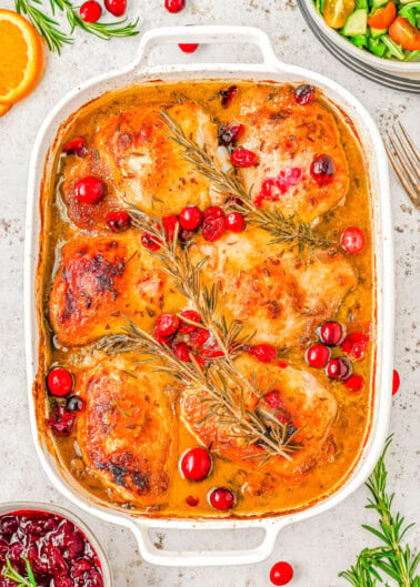 Baked chicken thighs in a casserole dish with cranberries and rosemary, garnished with orange slices.