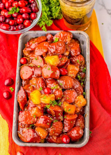 Rectangular dish with glazed sausage slices and pineapple chunks, garnished with parsley. A bowl of cranberries and a glass of iced drink are in the background, on a red cloth.