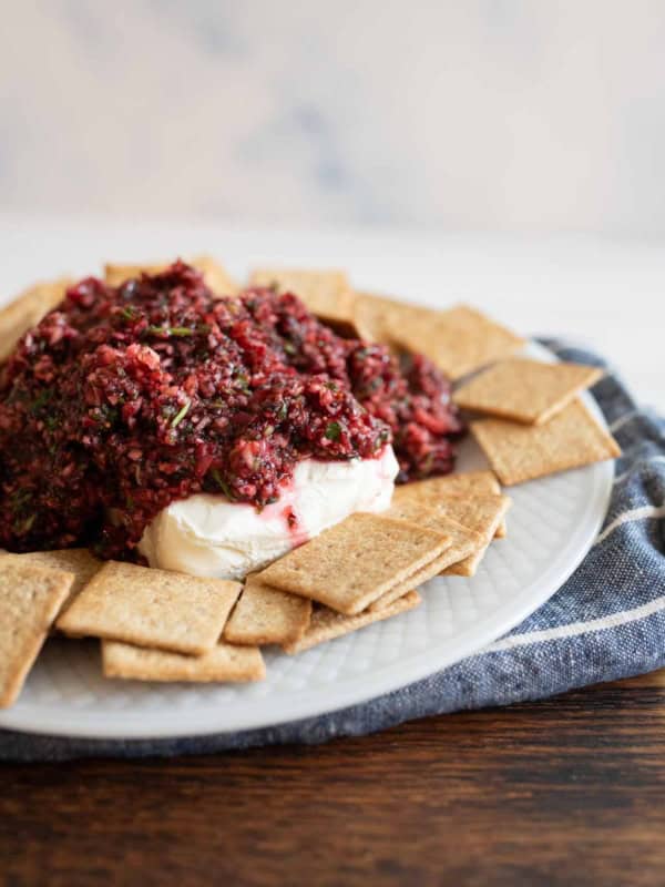 A plate topped with a mound of cranberry salsa on cream cheese, surrounded by square crackers. The setting is on a wooden table with a blue cloth underneath the plate.