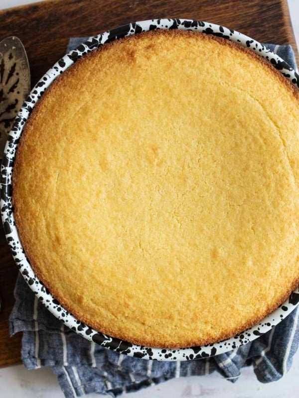 A freshly baked sweet cornbread in a round, black-and-white speckled ceramic dish sits on a wooden cutting board with two vintage serving utensils beside it. A striped cloth is partially under the dish, adding a rustic touch to this delightful recipe scene.