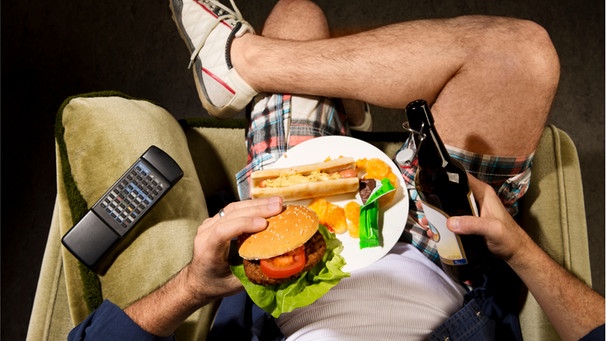 Ein Mann sitzt in einem Sessel mit einer Flasche Bier und Essen. - Gewohnheiten strukturieren den Tag, sie entlasten und belohnen uns. Unser Gehirn wäre ohne sie überfordert. Der Nachteil: Schlechte Angewohnheiten werden wir nur schwer wieder los. | Bild: Wochit/Getty Images