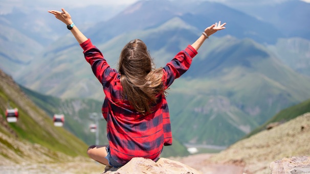 Glückliche Frau in den Bergen streckt die Hände in den Himmel. Resilient sein, Stress, Krisen und Schicksalsschläge überwinden - das wünschen sich viele. Welche Faktroren beeinflussen unsere Resilienz? Was macht uns stark und widerstandsfähig? | Bild: picture alliance / Zoonar | Svetlana