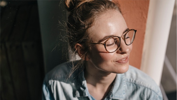 Eine Frau schaut aus dem Fenster raus, genießt die Sonnenstrahlen, die auf ihr Gesicht fallen. Der Urlaub ist vorbei und ihr seid gut erholt. Wie könnt ihr euch die Urlaubsstimmung und Entspannung im Alltag länger bewahren? Wir haben 6 Tipps für euch. | Bild: Wochit/Getty Images