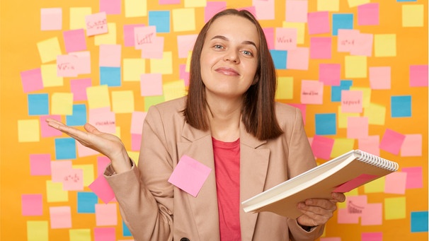 Eine junge Frau steht vor einer Wand mit vielen Post its, sie hat einen Schreibblock in der Hand. Habt ihr eine ständige To-do-Liste im Kopf, mit unsichtbaren Aufgaben, an die ihr denken müsst? Diese mentale Belastung nennt man auch Mental Load. Besonders Frauen und Mütter leiden darunter. Das könnt ihr dagegen tun ... | Bild: Wochit/Getty Images