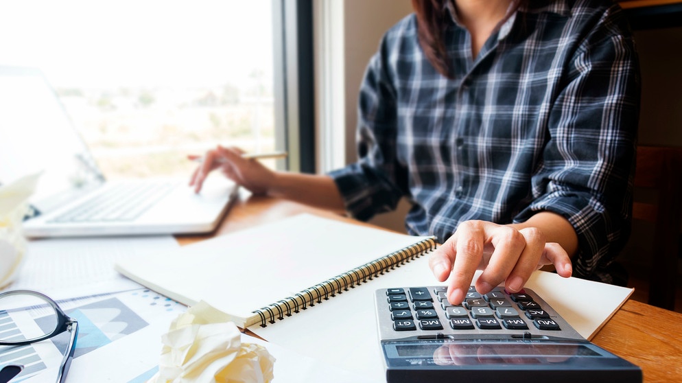 Frau rechnet mit einem Taschenrechner Kosten nach - vor ihr steh ein Laptop | Bild: mauritius images / Piyamas dulmunsumphun / Alamy / Alamy Stock Photos