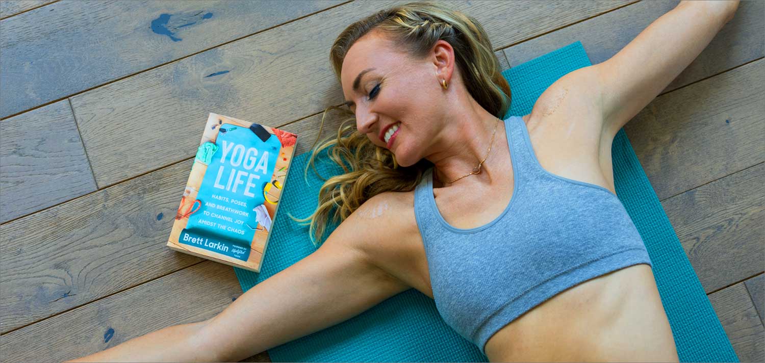 Yoga teacher lying in a supine twist on a blue yoga mat with the Yoga Life book 