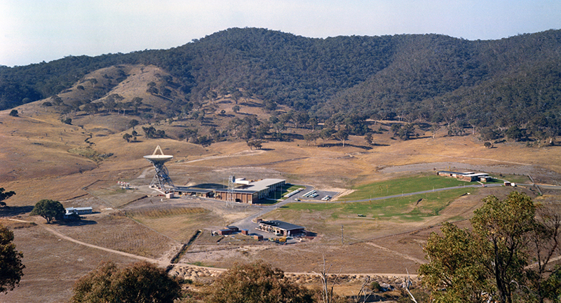 Hills covered in trees with a bare area in front with a couple of buildings, some roads, and one antenna.