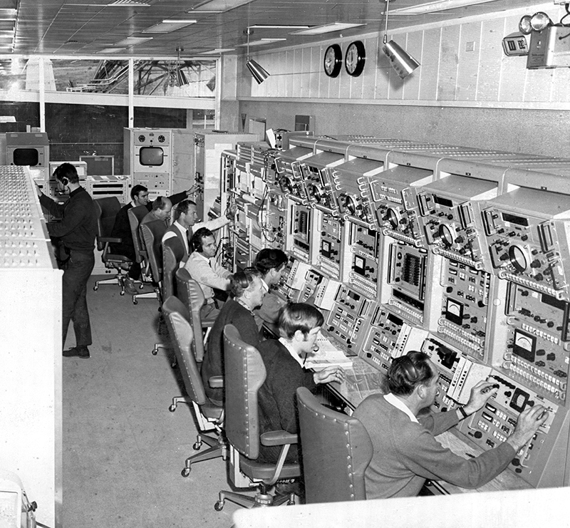 Black and white, eight men sitting at old very large computer consoles with one standing behind facing the other way.