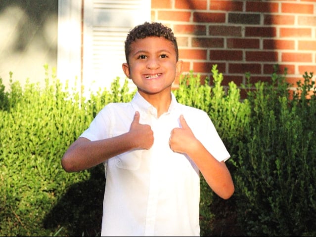 Eight-year-old Hezekiah, who has brown, curly hair, stands outdoors smiling and doing a thumbs up.