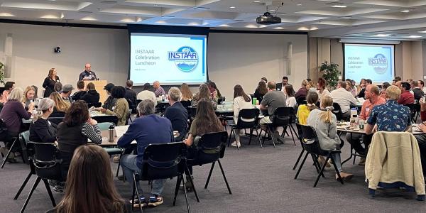 INSTAAR celebration luncheon May 8th 2024: A large room full of round tables packed with dozens of seated people having lunch and listening to opening remarks from Interim Director Nikki Lovenduski and Assistant Director of JEDI Julio Sepúlveda