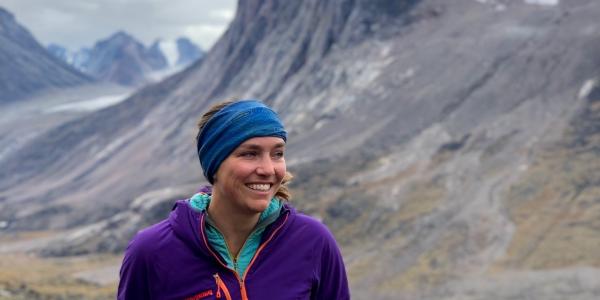 Sarah Crump, in purple coat, stands in front of a dramatic Baffin Island landscape of colorful tundra suddenly broken by towering cliffs.