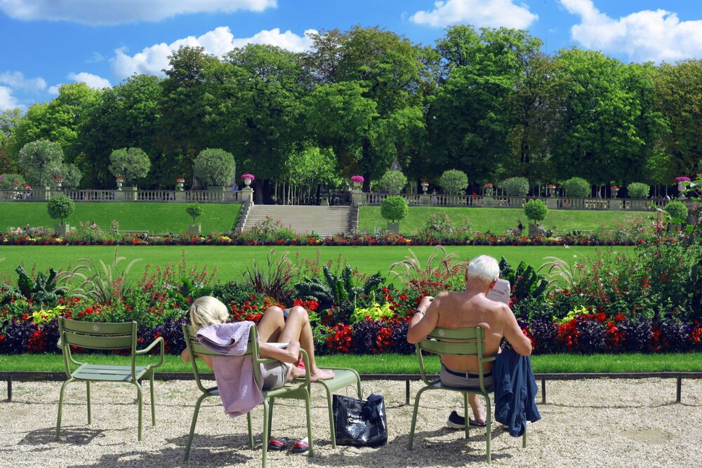 Two people sunbathing on chairs, looking over the well kept gardens with grass and flowers of the Jardin du Luxembourg.