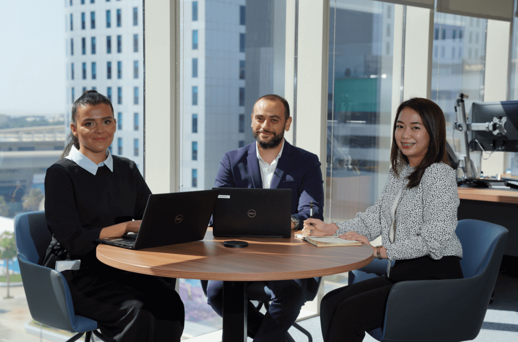 Three business professionals engaged in a meeting with laptops in a modern office with city view windows.