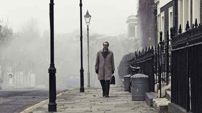 A man wearing a long coat and holding a bag walks on a sidewalk with street lamps