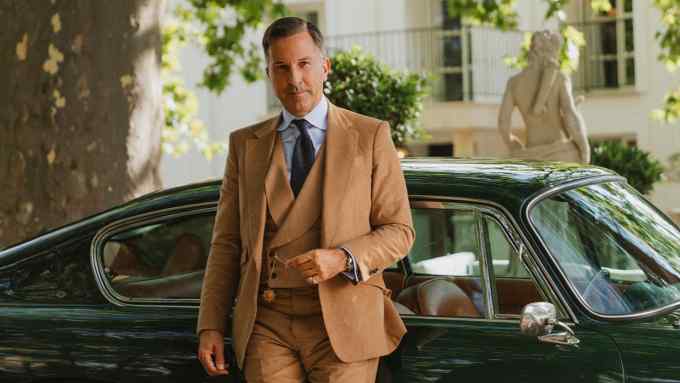 A man dressed in a stylish tan suit, standing in front of a classic green car. A small dog is seated beside him