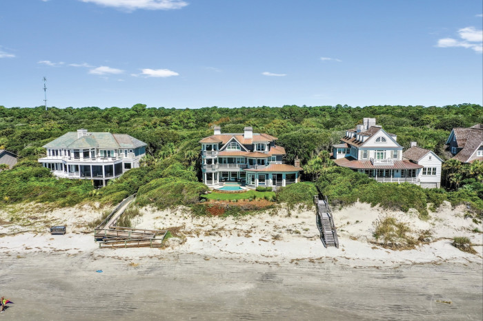 Aerial view of luxury beachfront homes nestled among lush greenery on a sandy beach, featuring a variety of architectural styles with multiple balconies and a private wooden walkway leading to the beach
