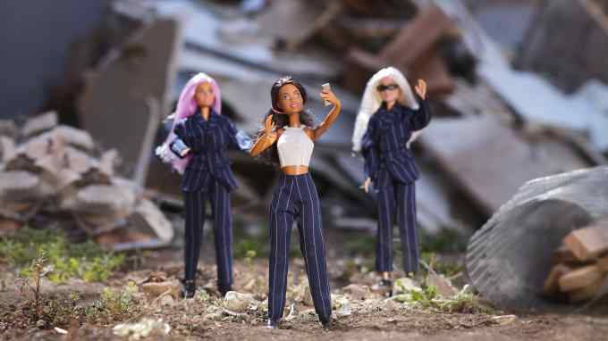 Three dolls stand in an outdoor setting with rubble and debris in the background. The central doll, with dark hair and a white crop top, poses for a selfie. The other two dolls, with pink and white hair respectively, wear dark pinstripe suits and stand behind her