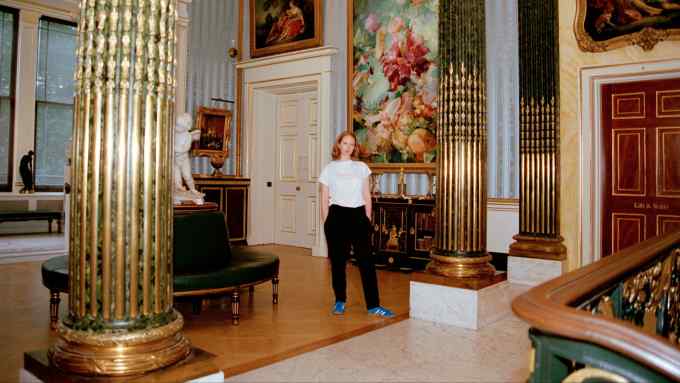 A woman wearing a white blouse and black trousers standing in front of a large multi-coloured abstract painting with a gold-coloured frame. Beneath is a black and gold ornate sideboard with gold-coloured ornaments on top. There are also ornate gold-coloured columns nearby