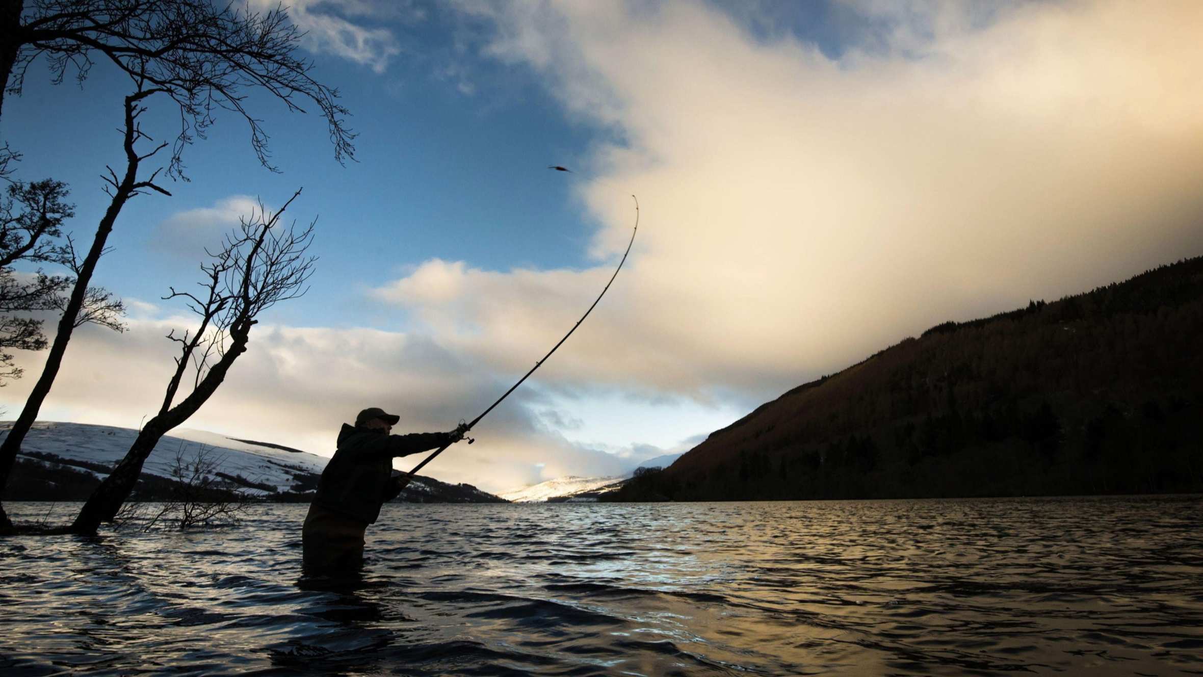 a fisherman in a river casting his rod with hill and dramatic clouds in background
