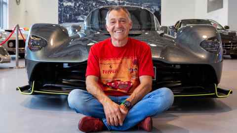 A man in a red Ferrari t-shirt sits cross-legged and smiling in front of a sleek, dark sports car in a showroom filled with other luxury cars