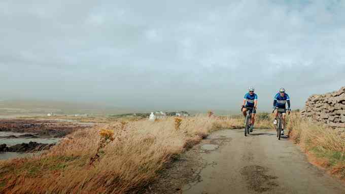 A front-on view of two cyclists in full sports cycling gear riding along a narrow hilltop country lane across moorland, with a drystone wall to their left