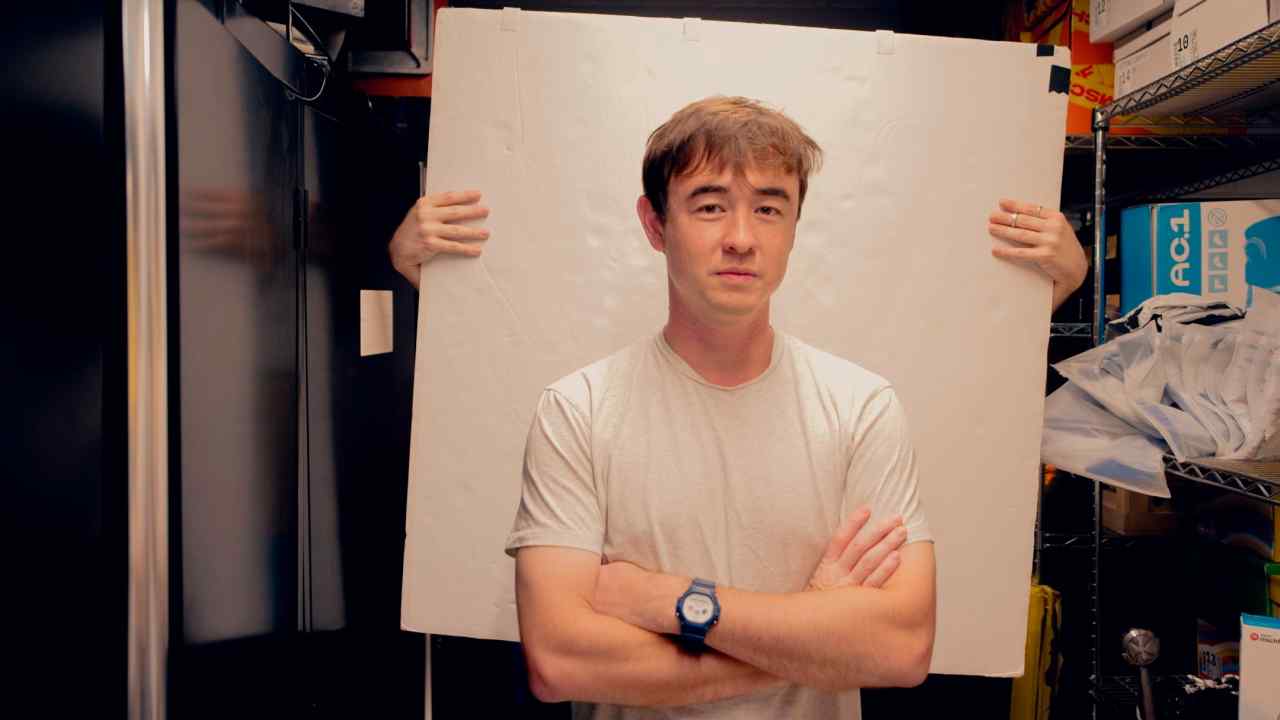 Man in white t-shirt in storeroom with person holding white board behind him