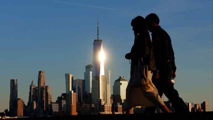 A young man and woman are walking at dusk. Behind them the skyscrapers of Manhattan glint in the setting sun