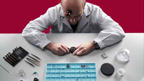 A professional watchmaker engaged in repairing a black wristwatch, using fine tools laid out beside him on a workstation with various small components sorted in a blue organizer under a bright work light
