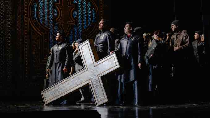 Three men looking stern in leather uniforms stand behind a large white cross tilted on the stage