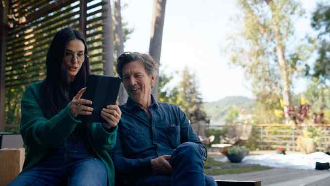 Demi Moore and Andrew McCarthy, dressed casually in jeans, sit side-by-side on a garden patio surrounded by trees looking at an electronic tablet