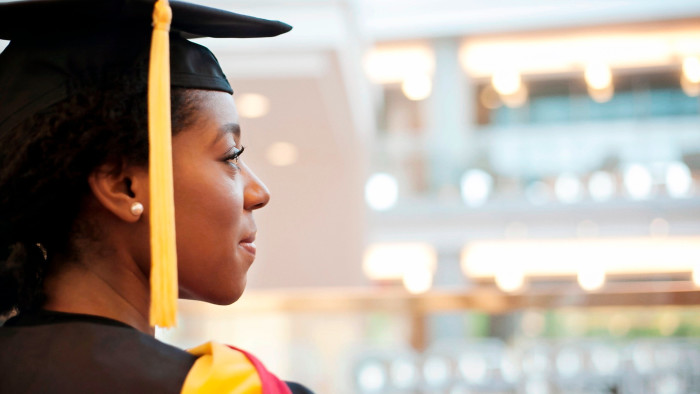 A close-up side profile of a young woman wearing a graduation cap and gown, looking ahead with a thoughtful expression. She is indoors with a brightly lit background