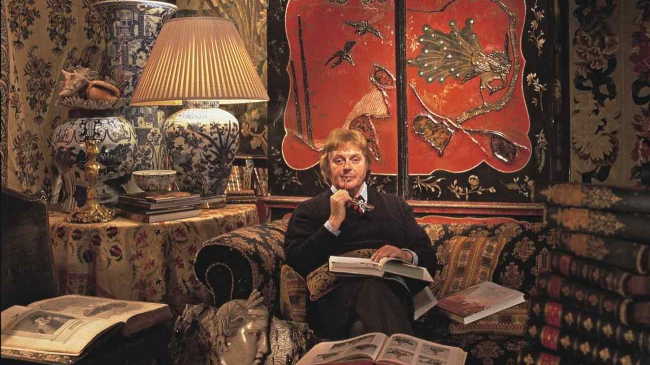 Geoffrey sits with books open in a room with tapestries, Chinese vases and lacquered artwork