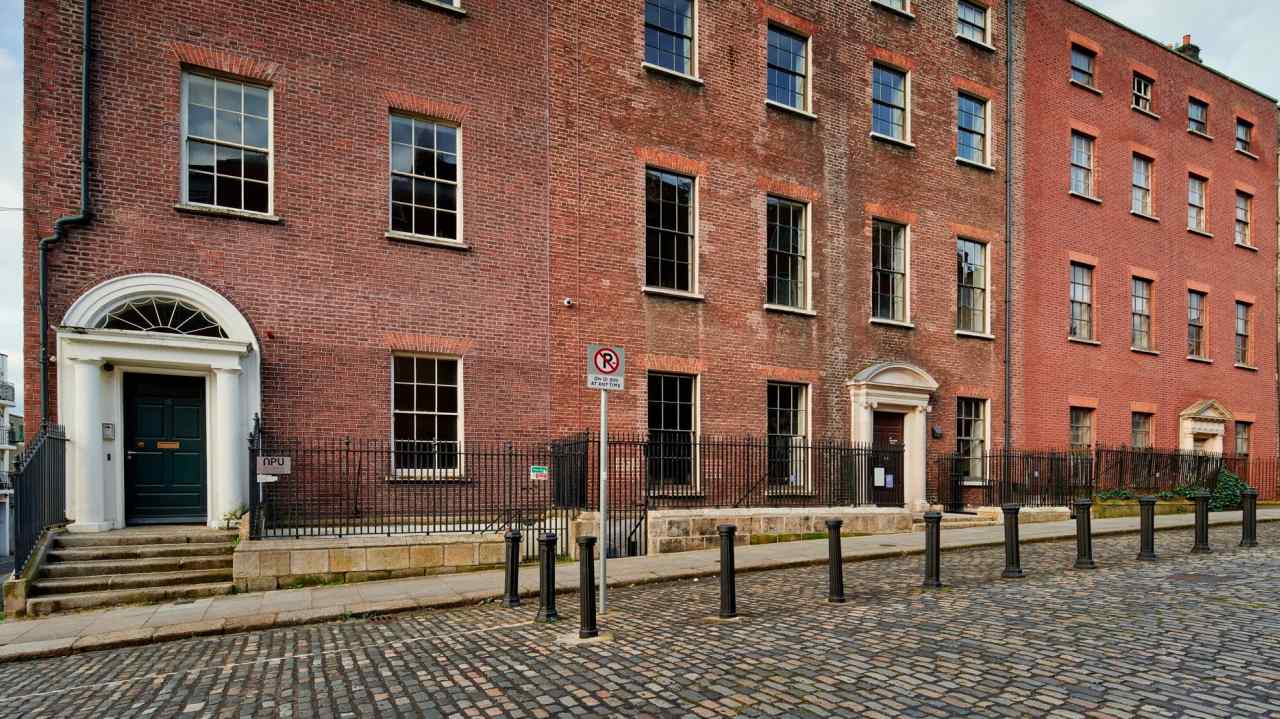 Georgian-style brick buildings with large sash windows and white-trimmed doorways. The cobblestone street in front is lined with black bollards, and a ‘No Parking’ sign is visible