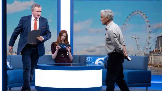A man in a suit stands next to a presenter’s desk on a TV set, talking to a man in shirt sleeves. A woman sits on the sofa behind the desk