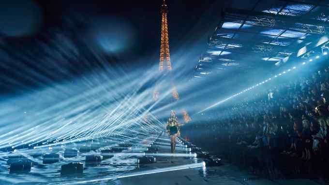 A model bathed in light walks along a runway at night, with the Eiffel Tower behind her