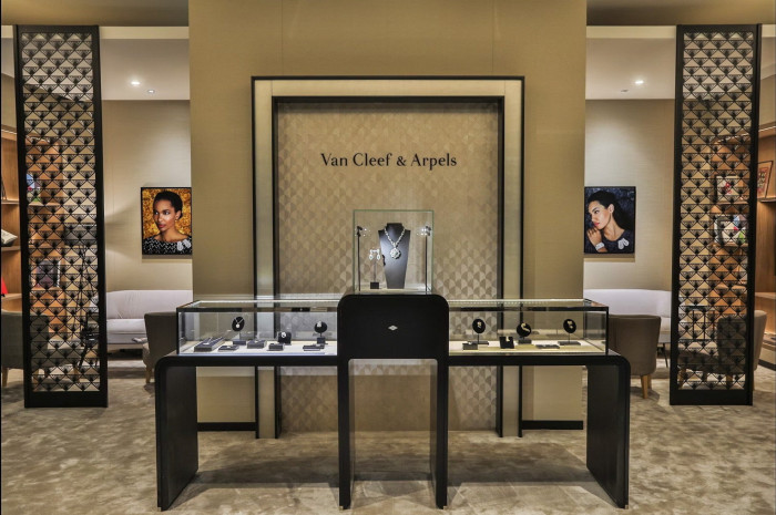A Van Cleef & Arpels exhibit showcasing a modern design with geometric patterns on the walls, a display table with jewelry pieces, and decorative portraits of models wearing the jewelry