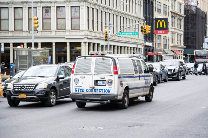 A New York City Department of Correction van driving on a city street with other vehicles around, including a McDonald’s restaurant in the background