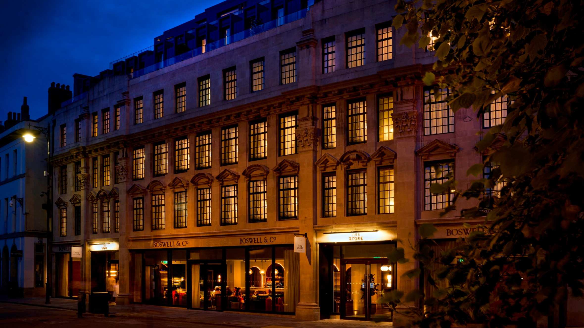 Four-storey stone building with an additional modern floor on top. All lit by internal lighting at dusk