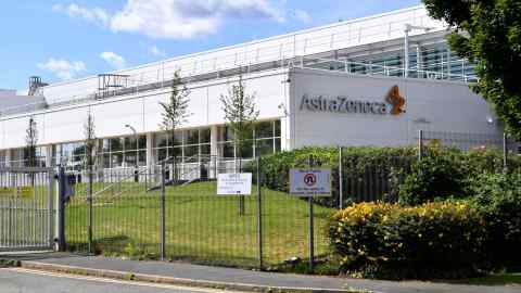 The exterior of the AstraZeneca factory in Speke, Liverpool, is shown on a sunny day. The factory building is white with the AstraZeneca logo prominently displayed. There is a fence with signs, including one that directs deliveries to a specific gate and another that prohibits U-turns for safety reasons. The area is landscaped with green grass, trees, and bushes