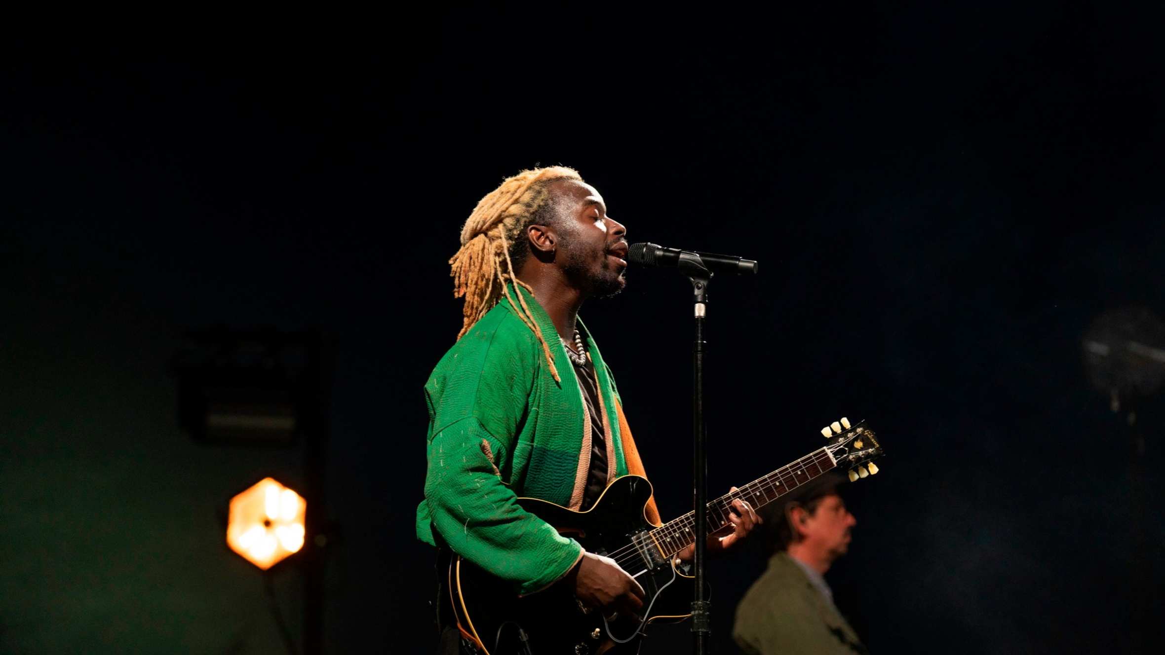 A man with blond locs and wearing a bright green jacket plays an electric guitar on stage while singing into microphone with his eyes closed