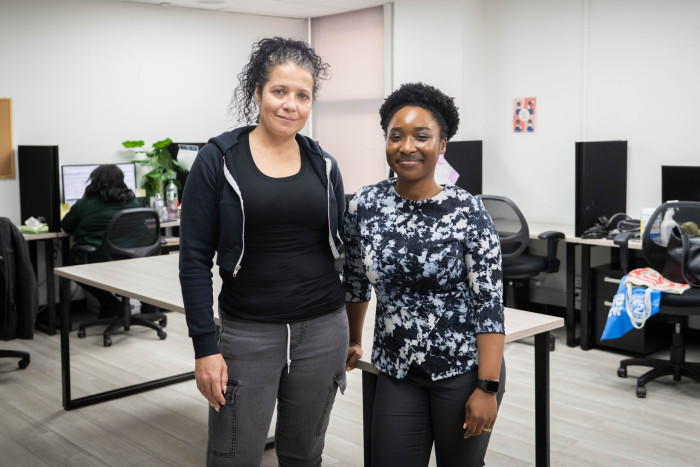 Two women smiling at the camera in an office setting, one Black woman in a floral blouse and another Caucasian woman in a black hoodie and gray pants, with computer workstations in the background