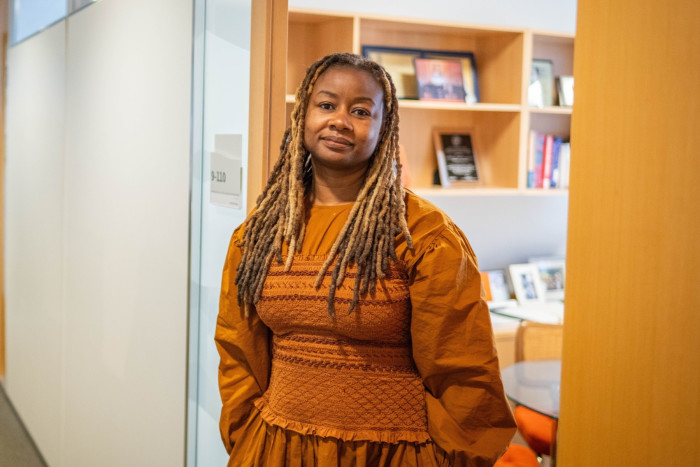 Portrait of a professional Black woman in a textured orange dress, smiling gently in an office environment with a modern glass door and a wooden bookcase in the background