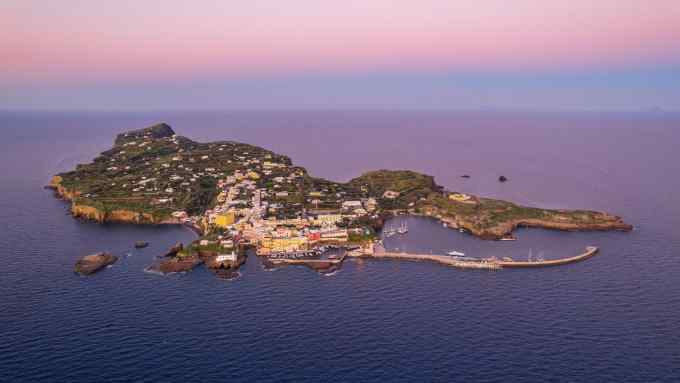 An aerial shot of an island just big enough to hold a hillside to the left, one village and a harbour to the right in the midst of a dark blue sea