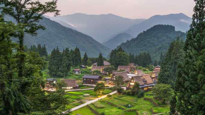 A lush green mountain valley at dusk with terraced fields and a scattering of houses with steep roofs, many thatched