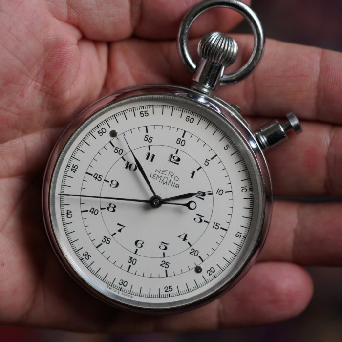A vintage-style stopwatch with a white dial and round metal case held in hand. The loop and crown are at the top. The pusher is at the north-east side of the face