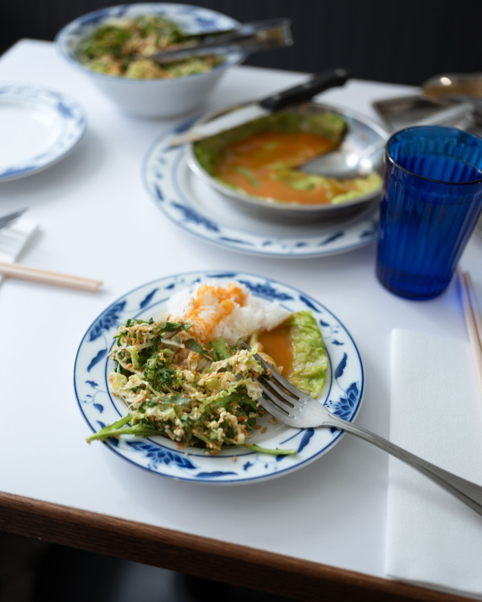 A small plate of stuffed Napa cabbage and chrysanthemum greens on a white-surfaced table, with another dish and a blue glass behind it