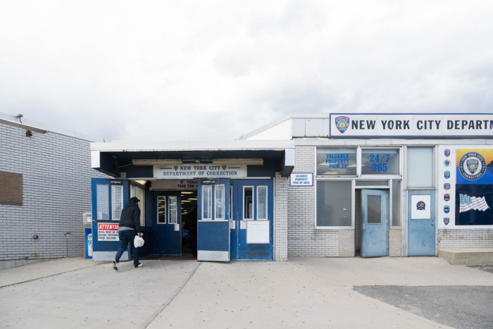 A man approaches the main entry of the New York City Department of Correction, marked by blue and white colors and clear signage, with a partly cloudy sky above