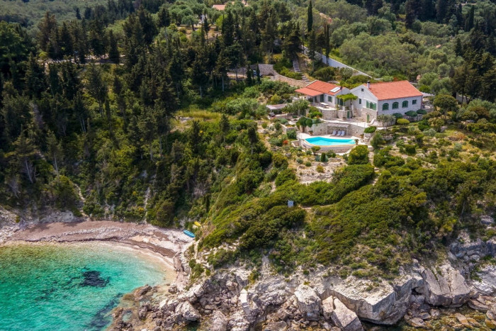 Aerial view of a secluded luxury villa with a red tile roof and swimming pool, nestled on a cliffside overlooking a clear turquoise sea, surrounded by dense green forest and terraced gardens