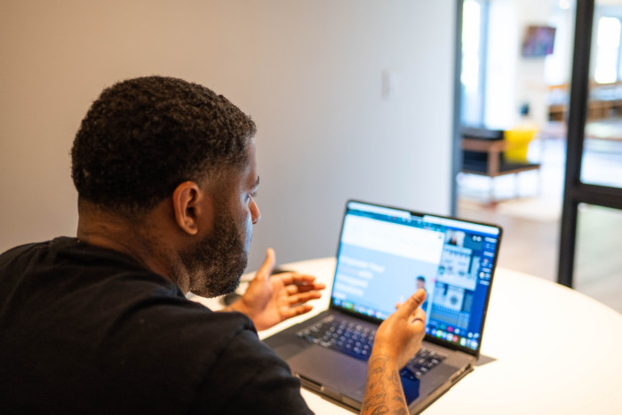 African-American male with tattoos on his arms engaging in a video call or presentation, using a laptop on a white table in a modern home office environment