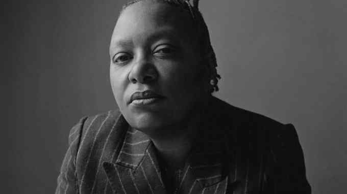 A black and white photo of Meshell Ndegeocello staring unsmiling directly at the camera, wearing a pin-striped blazer